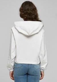 Felpa bianca accorciata con cappuccio a cordino, polsini elastici e una texture liscia, abbinata a jeans attillati azzurri.