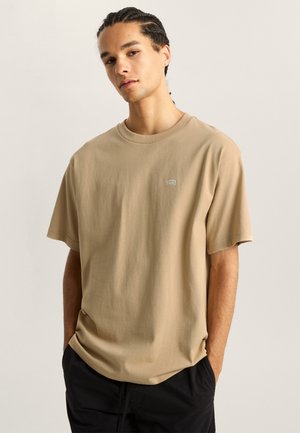 Light brown cotton t-shirt with a crew neck, short sleeves, and a small embroidered logo on the left chest. Casual fit and texture.