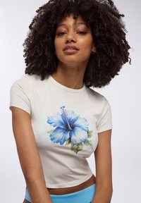 Crop top a maniche corte di colore crema con una stampa centrale di hibiscus blu e foglie verdi. Materiale in cotone morbido.