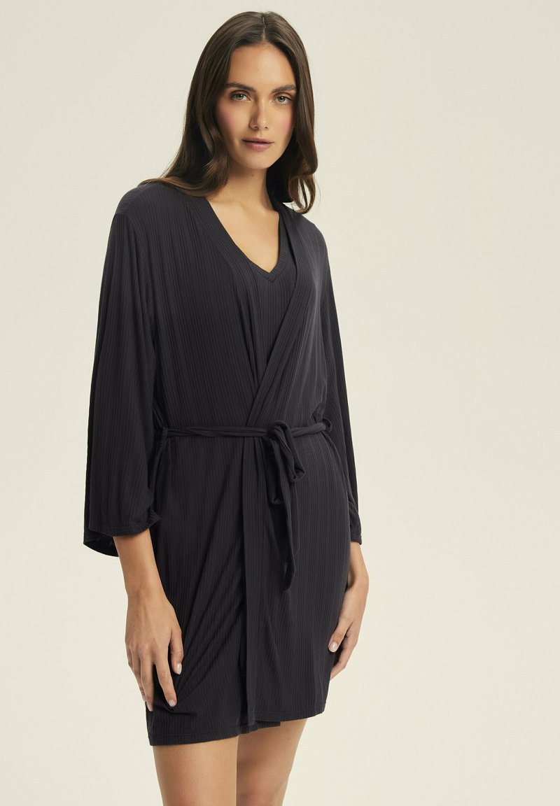 Czarna sukienka z prążkowanego materiału o kroju kopertowym, z dekoltem w kształcie V, długimi rękawami i paskiem w talii. Miękka tekstura, długość do kolan.