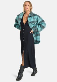 Oversized teal en grijs geruite jas over een zwarte maxi-jurk met knopen en een zijsplit, gecombineerd met zwarte enkellaarsjes.