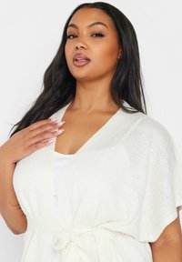 Vit, lätt cardigan med korta ärmar och knytning i midjan. Mjuk textur med en subtil lyster, som har en avslappnad passform och V-ringning.