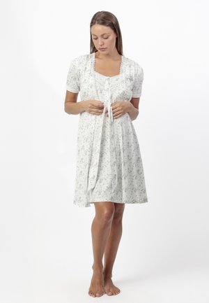 Joven mujer con camisón blanco floral hasta la rodilla, de pie descalza y mirando hacia abajo mientras sostiene los lazos delanteros de la prenda.