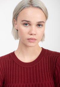 Jonge vrouw met kort blond haar en een lichte huid, gekleed in een ribbelige bordeauxrode trui, kijkt recht vooruit met een neutrale uitdrukking tegen een witte achtergrond.