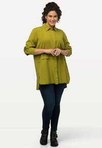 Camisa de botones de color verde oliva con mangas largas y un diseño en capas, combinada con vaqueros de color azul oscuro y botas negras hasta el tobillo.