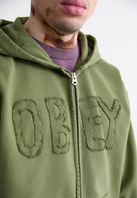 Olive green zip-up hoodie with large, stitched “OBEY” lettering on the front. Features a hood and silver zipper detail.