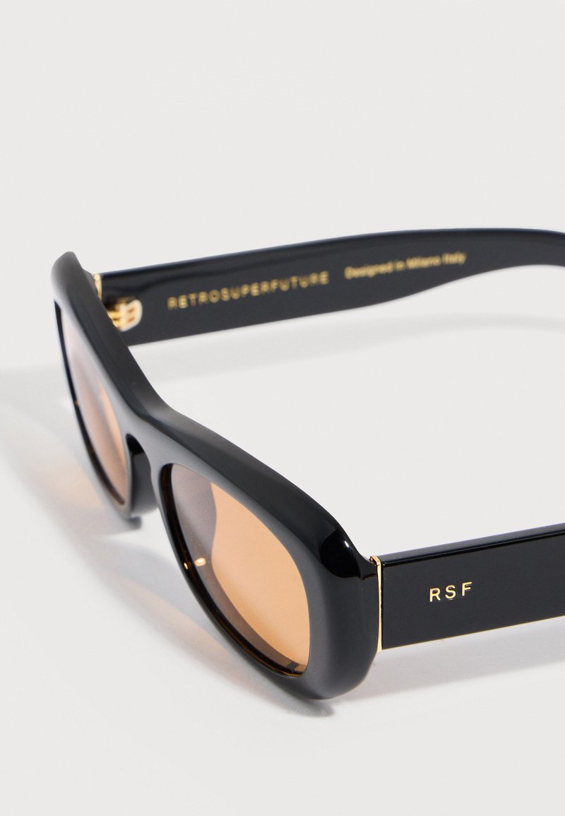 Gafas de sol rectangulares negras con lentes marrones, detalles dorados y letras "RSF" en la patilla, anguladas sobre una superficie blanca.