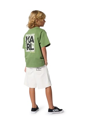 Enfant aux cheveux blonds portant un T-shirt vert Karl Lagerfeld, un short blanc avec logo, et des baskets noires avec le texte "KARL", debout de côté.