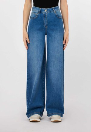 Donna che indossa jeans a vita alta a gamba larga blu e sneakers bianche, in piedi con le mani rilassate ai lati contro uno sfondo semplice.