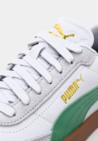 Baltie sporta apavi ar pelēkiem zamšādas akcentiem, zaļu sānu joslu, zelta logotipu un plakaniem baltajiem šņorēm. Iezīmējas ar strukturētu augšējo daļu un brūnu gumijas zoli.