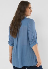 Blusa blu a maniche lunghe in tessuto leggero con polsini arrotolati, una spalla posteriore e una vestibilità rilassata. Superficie testurizzata, accenti minimi.