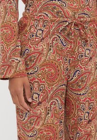 Červená látka s paisley vzorem a zlatými a modrými akcenty, s elastickým pasem a dlouhými rukávy. Materiál vypadá hladce a má detailní design.