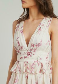 Kwiatowa sukienka z lekkiego, kremowego materiału z różowymi wzorami kwiatowymi, głębokim dekoltem w kształcie litery V, plisowanym korpusem oraz warstwowymi falbanami na talii.