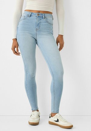 Lichtblauwe skinny jeans met een hoge taille, voorzien van subtiele vervaagdetails en een koperen knop. Gecombineerd met wit en zwarte sneakers.