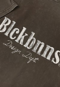 Bruine stof met gedurfde, verweerde witte tekst die "Blckbnn's Design Dept." zegt. Soepele textuur met een lichte glans.