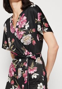 Mono floral de tela negra con flores en tonos rosa y blanco, escote en V, mangas cortas y un cinturón atado para un ajuste ceñido.