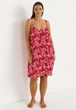 Kobieta stoi boso przy białej ścianie, nosząc luźną czerwoną sukienkę bez rękawów z białymi wzorami kwiatowymi.