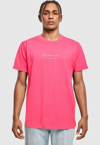 Camiseta de algodón rosa con mangas cortas, que presenta texto en árabe y la frase en inglés "Ámate a ti mismo primero", centrada en el pecho.