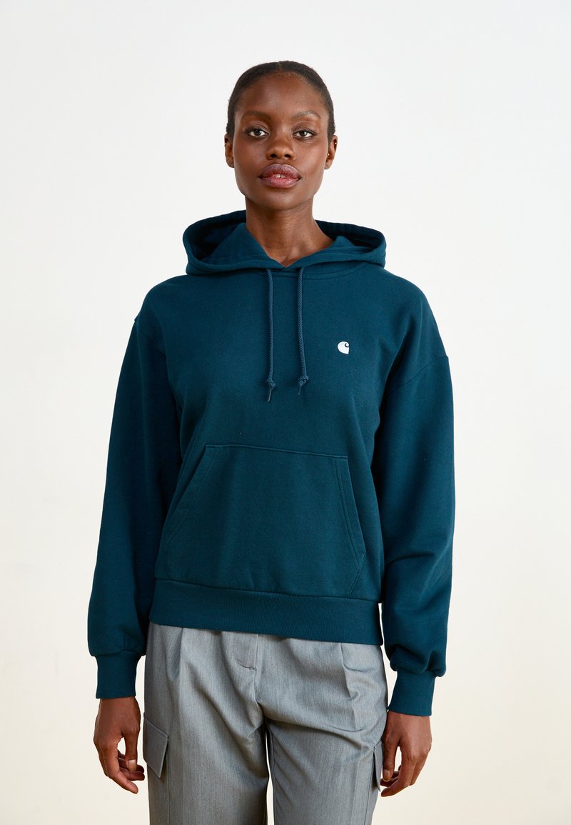 Dark teal hoodie made of soft fabric, featuring a front pocket, drawstring hood, and a small white logo on the left chest. Paired with gray pants.