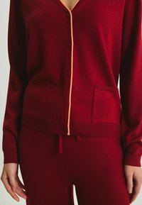 Cardigan en tricot rouge avec un col en V, des finitions orange et une fermeture à boutons. Il possède deux poches avant et une taille côtelée. Texture lisse.