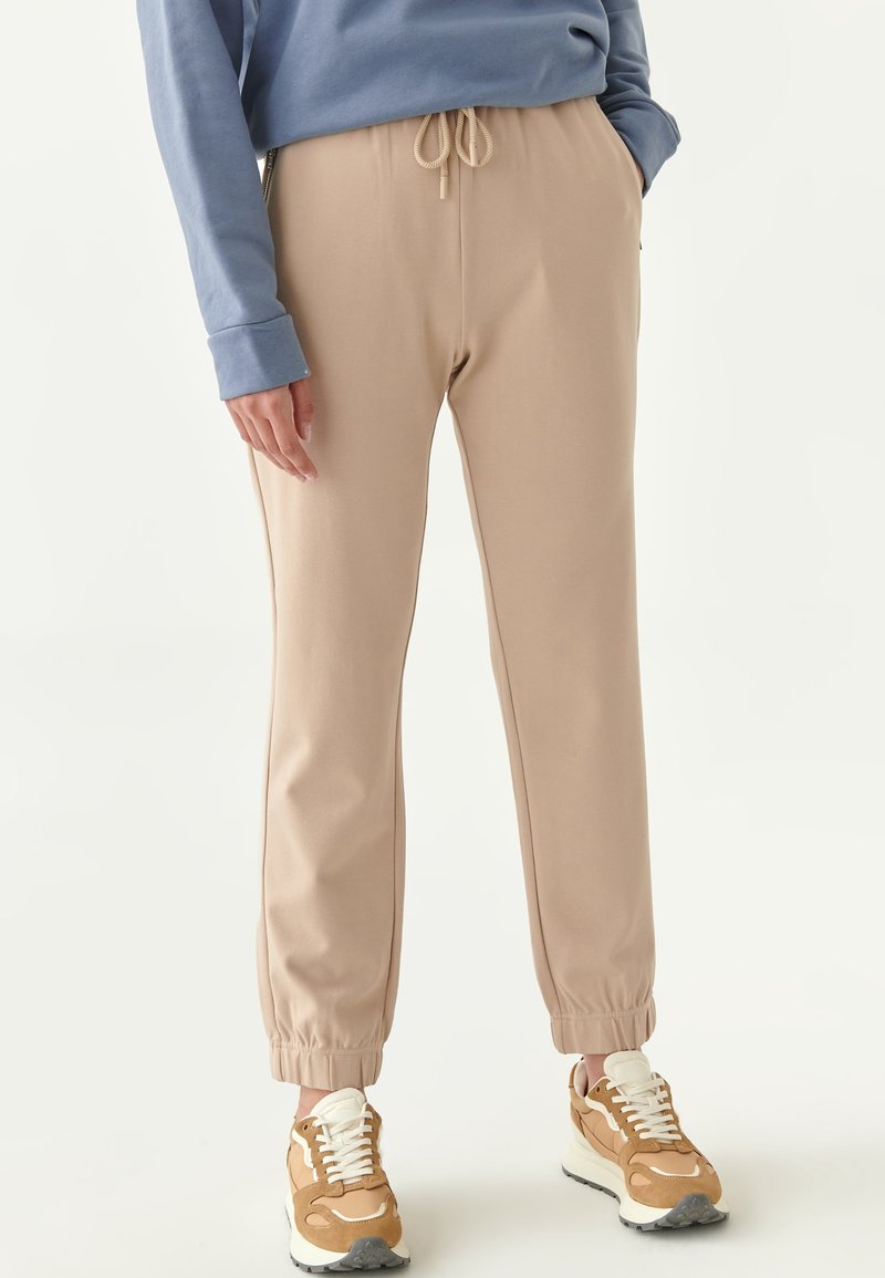 Beige Jogginghosen mit einem elastischen Bund und Ärmelbündchen, aus weichem Material. Kombiniert mit beige-weißen Sneakern für einen lässigen Look.