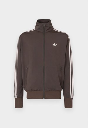 Brūns rāvējslēdzēja sporta žakete ar augstu apkakli, baltām sānu svītrām un logotipu krūtīs. Aprīkota ar divām rāvējslēdzēja kabatām priekšpusē un ribota apdare aprocēs.