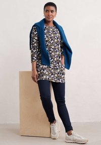 Kwiatowa tunika w oliwkowym kolorze z niebieskimi i białymi wzorami, zestawiona z granatowymi skinny jeansami i białymi trampkami, uzupełniona niebieskim swetrem.