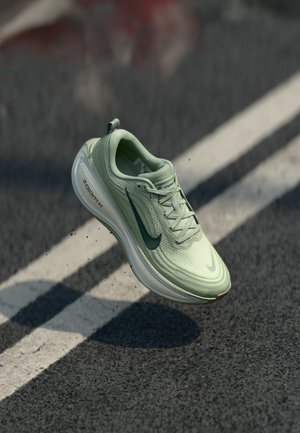 Lichtgroene Nike hardloopschoen met zwarte swoosh zweeft in de lucht boven een asfaltweg met witte lijnmarkeringen.