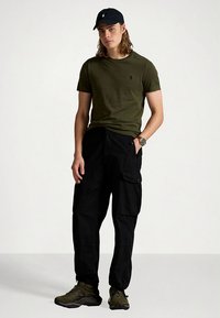 T-shirt verde-oliva, calças cargo pretas com bolsos, boné preto e ténis verde-escuro. Design simples com ajuste descontraído e cores neutras.