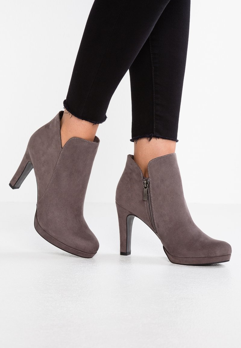 Bottes basses en daim gris avec fermetures éclair latérales et talons hauts, portées avec un pantalon noir à ourlet effiloché sur un fond de sol blanc.