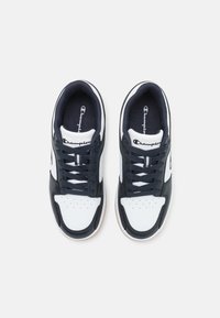 Chaussures de sport blanches et bleu marine avec une tige en cuir, bout rond, boîte à orteils perforée et semelle en caoutchouc. Logo Champion sur la languette.