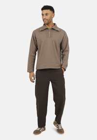 Pullover marrone con colletto a zip e texture a coste, abbinato a pantaloni marroni scuri e sneakers grigie con strisce bianche.