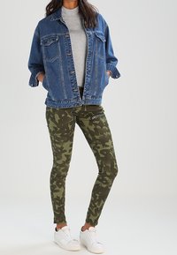 Jeansjacka i blått med två bröstfickor, grå t-shirt, gröna skinny-byxor med kamouflagemönster, och vita sneakers. Avslappnad outfit.