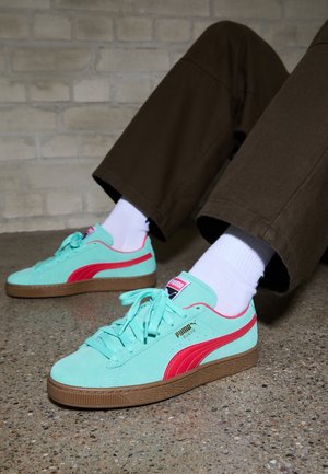 Personne portant des baskets en daim turquoise Puma avec une bande rouge, des chaussettes blanches et un pantalon marron, assise sur un sol tacheté contre un mur en briques.