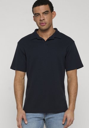 Mand med kort, krøllet hår, iført en sort kortærmet polo og lyseblå jeans, stående foran en ensfarvet baggrund.