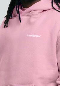 Růžová mikina s kapucí vyrobená z měkkého materiálu, s malým bílým logem na hrudi a volným střihem.