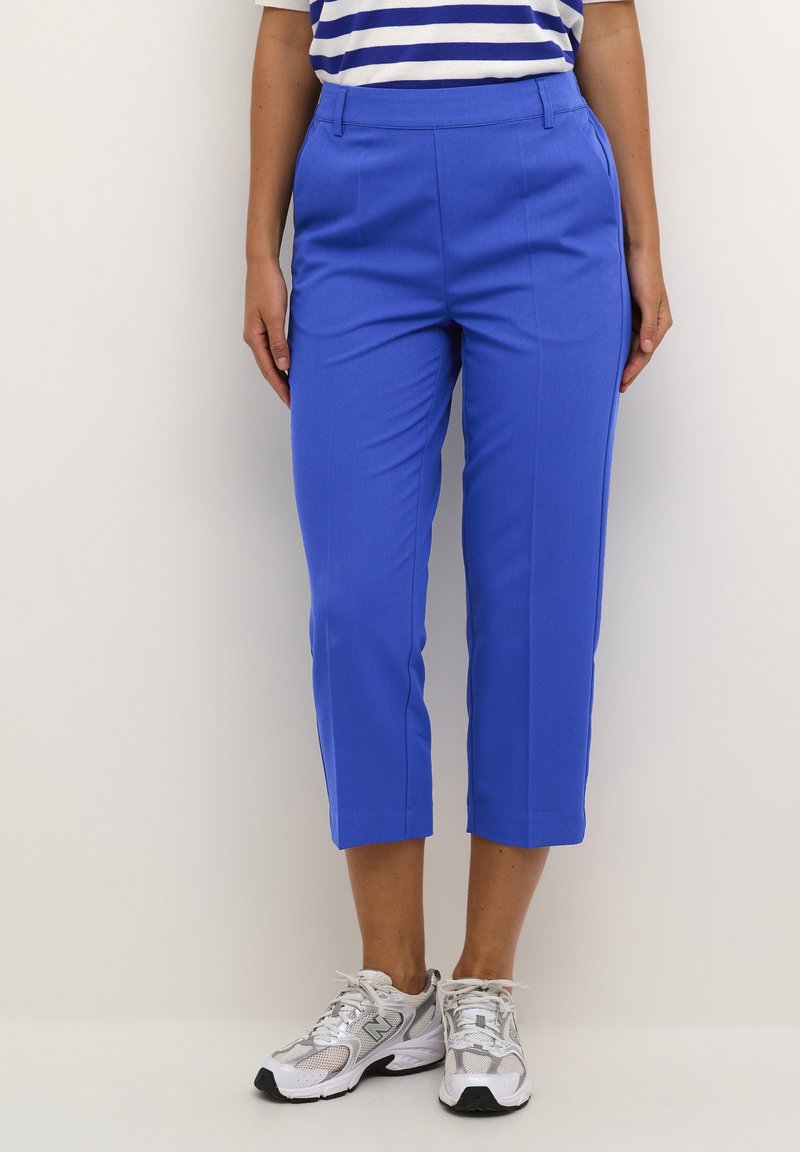 Gekürzte blaue Hosen aus glattem Stoff, mit einem schmalen Taillenbund und seitlichen Taschen, kombiniert mit weißen und grauen Turnschuhen.