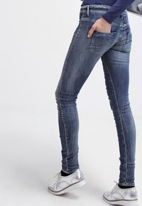Jean skinny en denim bleu foncé avec un délavage, arborant un design à cinq poches. Associé à des chaussures à lacets en argent métallique.