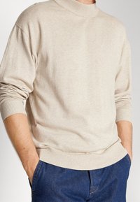 Mann in einem hellbeigen, langärmeligen Strickpullover mit gerippten Bündchen und Saum, Hände in den Taschen einer blauen Jeans, vor einem neutralen Hintergrund.