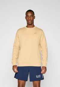 Lys beige genser med rund hals og ribbestrikkede mansjetter. Har et lite, diskret logo. Kombinert med mørk marineblå shorts. Jevn tekstur.
