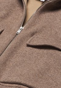 Brun zip-up tröja med en strukturerad yta, utrustad med två framfickor och ett mjukt innerfoder i en ljusare tan-färg. Silverfärgad dragkedja.