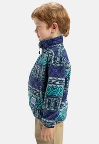 Veste en polaire avec motif géométrique en bleu, violet et turquoise. Col élevé, fermeture éclair à l'avant et poches. Texture douce, coupe ajustée.