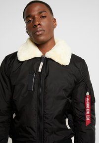 Chaqueta bomber negra con un cuello de piel sintética blanca, cierre frontal y múltiples bolsillos. Presenta una etiqueta roja que dice "RETIRAR ANTES DEL VUELO."