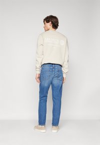 Beez ž sweatshirt, mille seljal on tekst "LES DEUX COMMUNITY", koos siniste teksapükstega. Mudel kannab beeže tossusid.