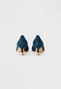 Une paire de chaussures à talons kitten en daim bleu foncé avec des talons aiguilles dorés, vues de l'arrière sur fond blanc.