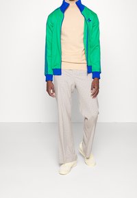 Vihreä ja sininen vetoketjutakki, beigenvärinen neulepaita, vaaleanruskeat ruutuhousut ja vaaleankeltaiset tennarit. Kangas on sileää ja puhdasta.