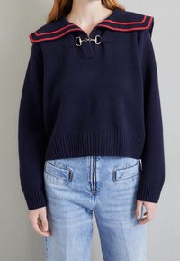 Marinblå tröja med en röd- och blårandig sjömanskrage, med stängning framtill med ögla och ribbad fåll, kombinerad med ljusblå jeans.