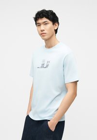 Νέος άνδρας με κοντά σγουρά μαύρα μαλλιά, φορώντας ανοιχτό μπλε κοντομάνικο μπλουζάκι με λογότυπο και σκούρο παντελόνι, στέκεται με τα χέρια στις τσέπες.