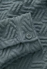 Texturovaná tmavě šedá tkanina s geometrickým vzorem, s detaily knoflíku na manžetě, ukazující jemný pletený vzhled.