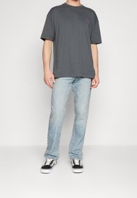 Harmaa puuvillapaita, jossa on löysä malli, purppura logo rinnassa, yhdistettynä vaaleansinisiin farkkuihin ja musta-valkoisiin tennareihin.